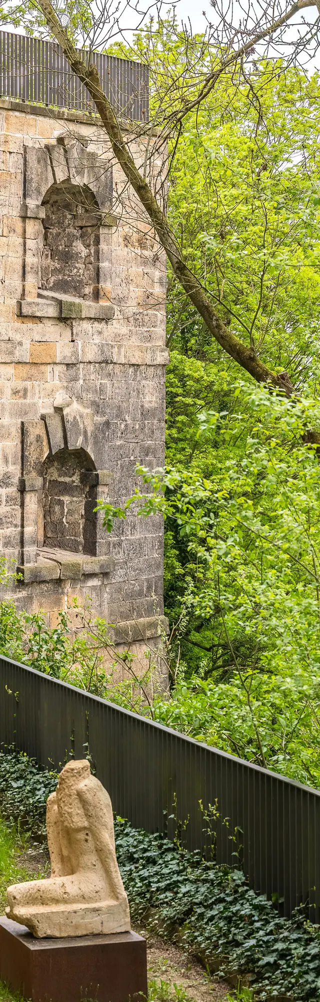 Alte Steinmauer des Schloss Sonnenstein mit Efeu bewachsen, umgeben von Bäumen; im Vordergrund stehen mehrere antike Skulpturen auf einer grünen Wiese.