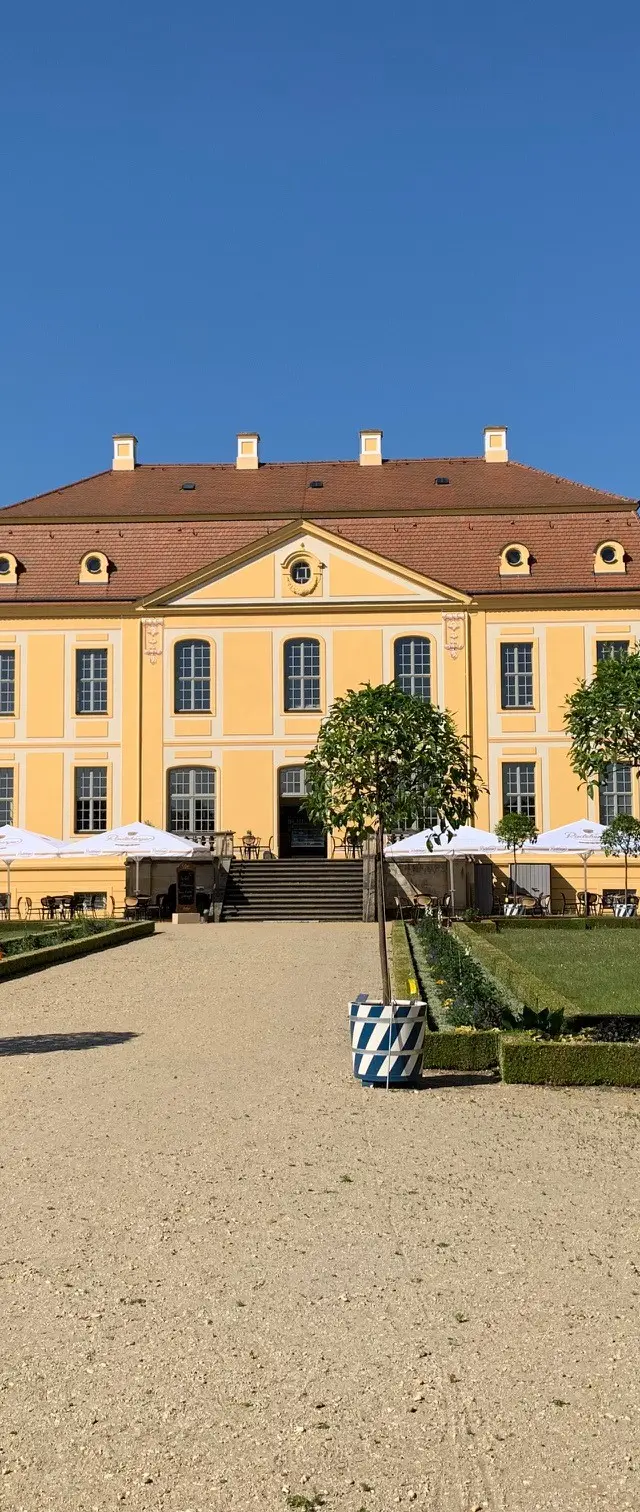 Gelbes Barockgebäude des Barockgarten Großsedlitz mit rotem Dach, umgeben von gepflegtem Garten und Bäumen in blau-weißen Töpfen, unter klarem blauem Himmel.