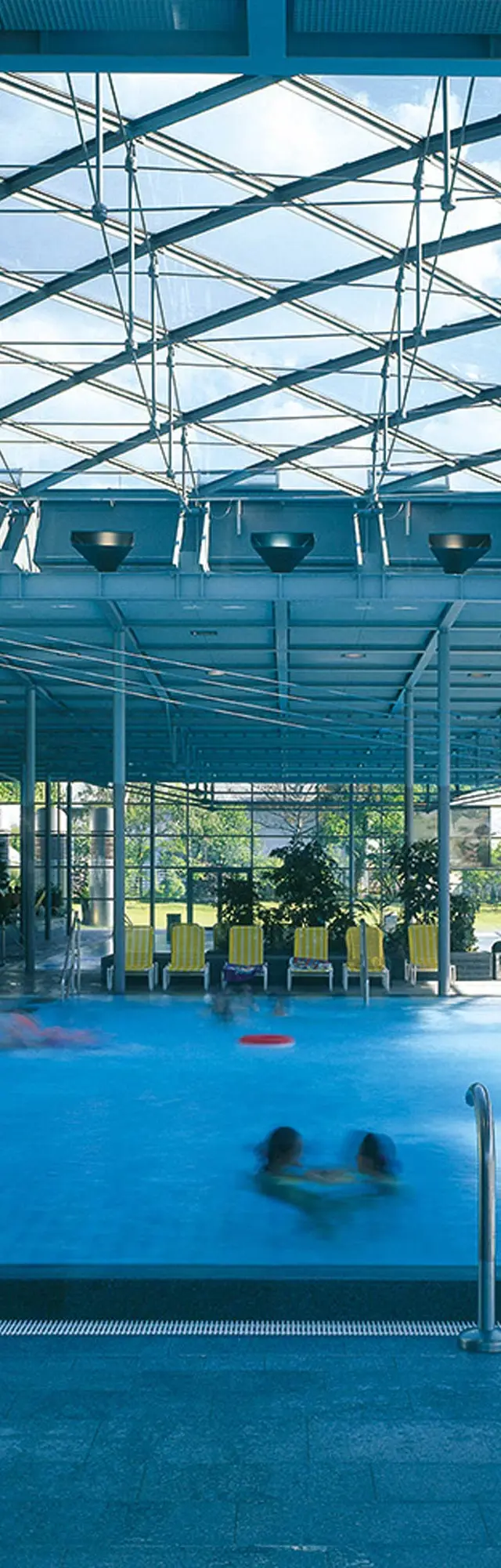 Geibeltbad Pirna: Modernes Hallenbad mit Glasdach, blauem Wasser und mehreren schwimmenden Personen; helle, luftige Atmosphäre mit Pflanzen und Liegestühlen.